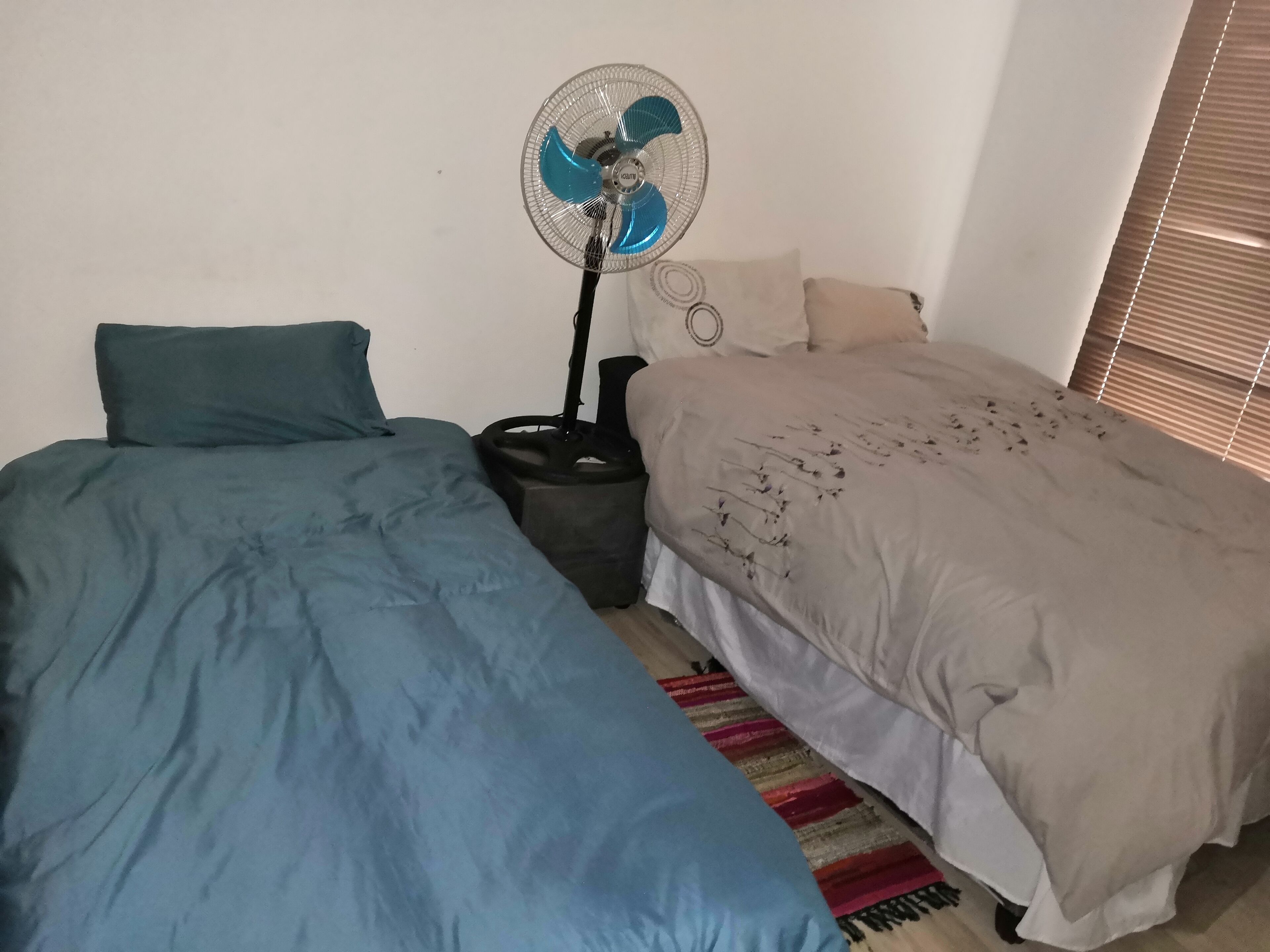 Ein Schlafzimmer mit zwei Betten, einem Standventilator und einem Fenster mit Jalousien.