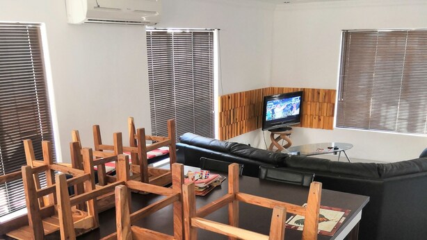A dining area with a wooden table and chairs, a television on a stand, and a window with blinds. - Family room (Khomasdal)
