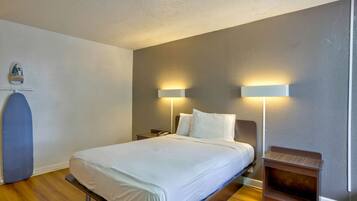 Un lit bien fait dans une chambre d’hôtel, avec deux lampes de chevet, une petite table de nuit en bois et une planche à repasser fixée au mur.