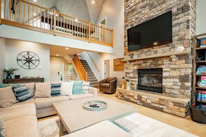 A spacious living area with a stone fireplace, a large flat-screen TV, a wooden staircase, and a cozy seating area.