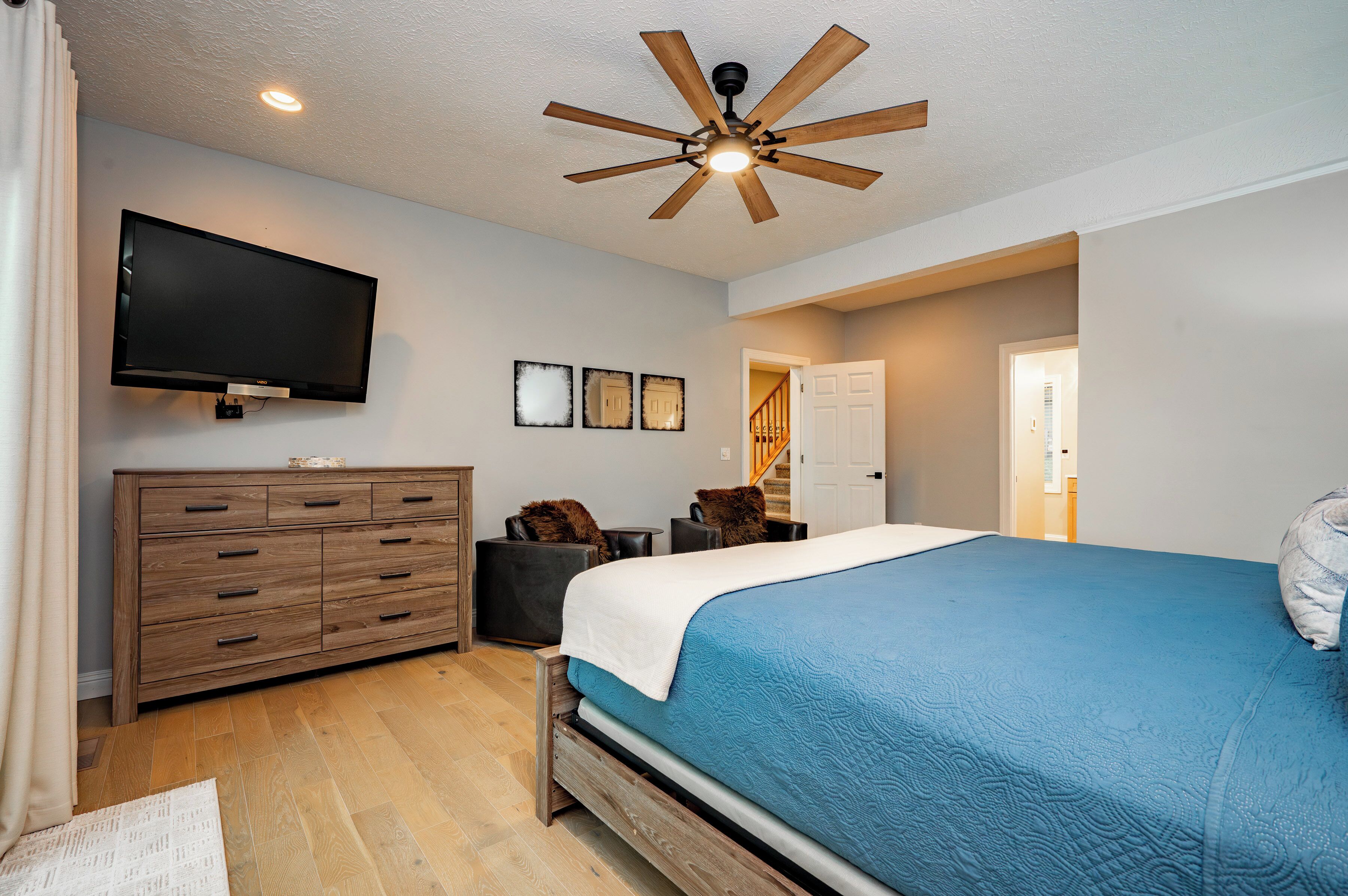 A bedroom with a bed, a TV, a ceiling fan, a dresser, and a door leading to another room.