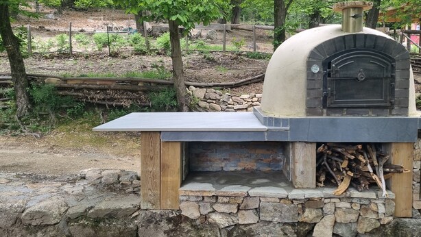 Un horno exterior de piedra y madera con tapa plana, rodeado de un entorno rústico con árboles y un muro de piedra.