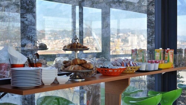Een ontbijtbuffet met croissants, gebak en vruchtensappen op een houten tafel, met een stadszicht door grote ramen.