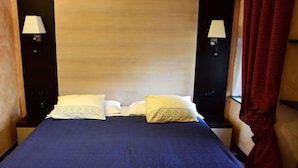 Een hotelkamer met een houten hoofdeinde, twee bedlampen, een bed met een blauwe plaid en een rood gordijn.