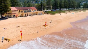 A beach with a lifeguard tower, a building with a red roof, and a sandy shore. - Holiday on the Waterfront in Pittwater -Beach, Swim, Kayak, Boat, Fish and More! (Newport)
