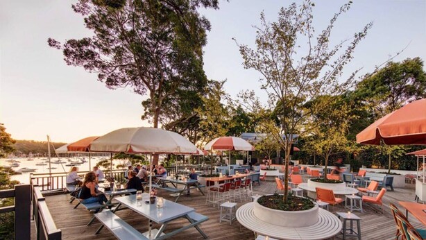 An outdoor dining area with tables and chairs, surrounded by trees and overlooking a body of water. - Holiday on the Waterfront in Pittwater -Beach, Swim, Kayak, Boat, Fish and More! (Newport)
