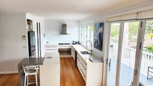 A modern kitchen with a central island, a built-in oven, and a large window overlooking a deck. - Holiday on the Waterfront in Pittwater -Beach, Swim, Kayak, Boat, Fish and More! (Newport)