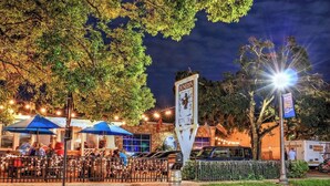 Un espace extérieur aménagé pour les repas en soirée, avec des tables et des parasols, une enseigne indiquant l’emplacement de la brasserie de Dunedin, et une vue sur la rue.
