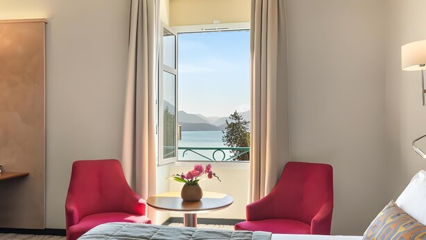 A hotel room with a bed, two red chairs, a small table with a vase, and a view of a lake and mountains. - Impérial Palace (Annecy)