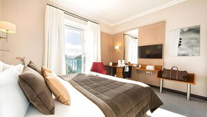 A hotel room with a large bed, a desk, a chair, a mirror, and a view of the outdoors. - Impérial Palace (Annecy)