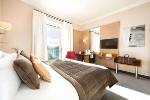 A hotel room with a large bed, a desk, a chair, a mirror, and a view of the outdoors. - Impérial Palace (Annecy)
