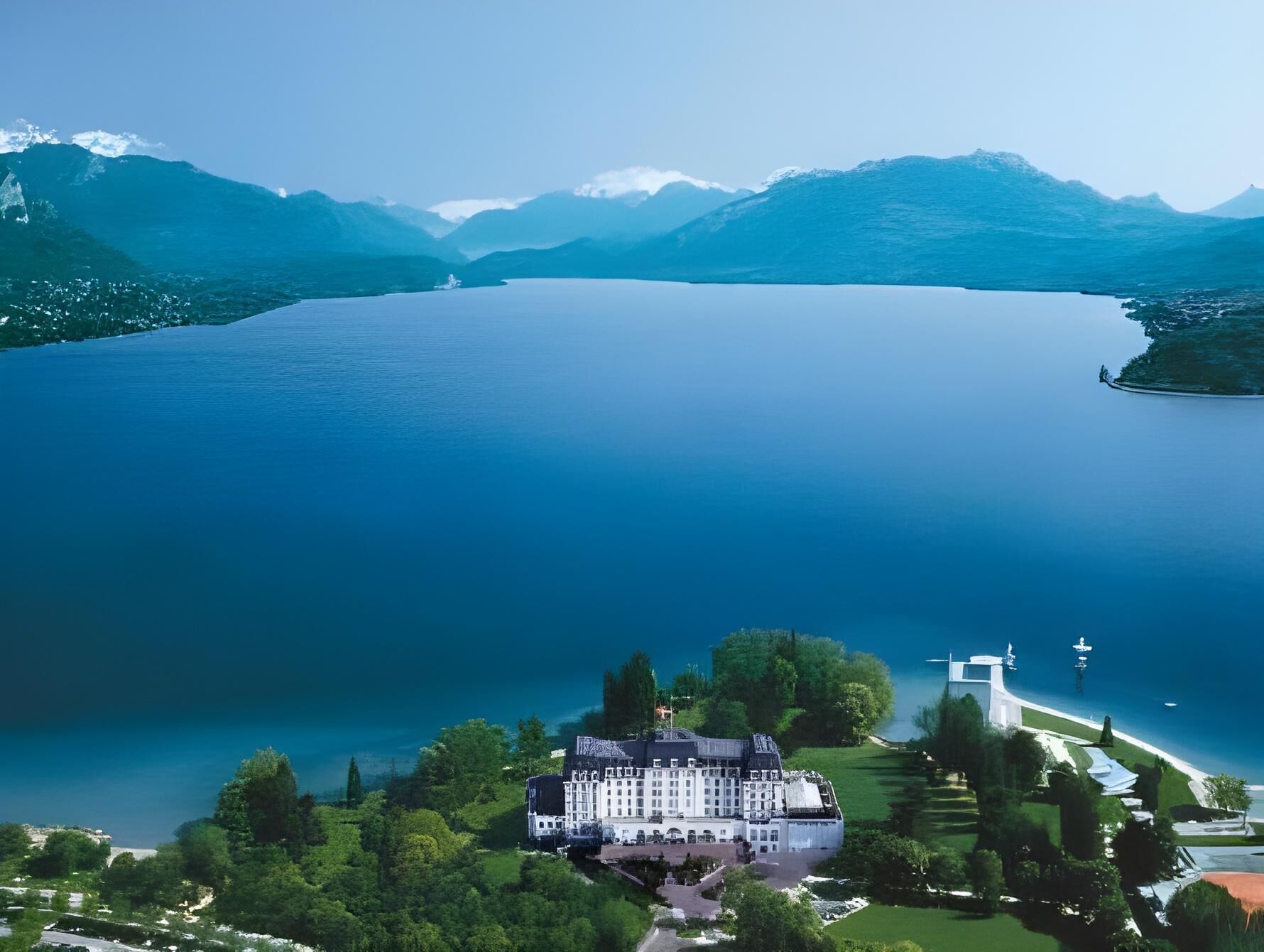 A large, white building with multiple floors, surrounded by lush greenery and a lake.