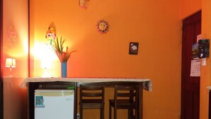 A compact kitchen with a refrigerator, two wooden chairs, a table with a white tablecloth, and a wall adorned with a sun decoration and a framed picture. - Weekly Rental Beach Sunny , Party Town Costa Rica (Coco Beach)