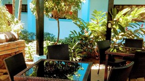 An outdoor seating area with wicker furniture and a glass-top table, surrounded by various plants and a blue wall in the background. - Weekly Rental Beach Sunny , Party Town Costa Rica (Coco Beach)