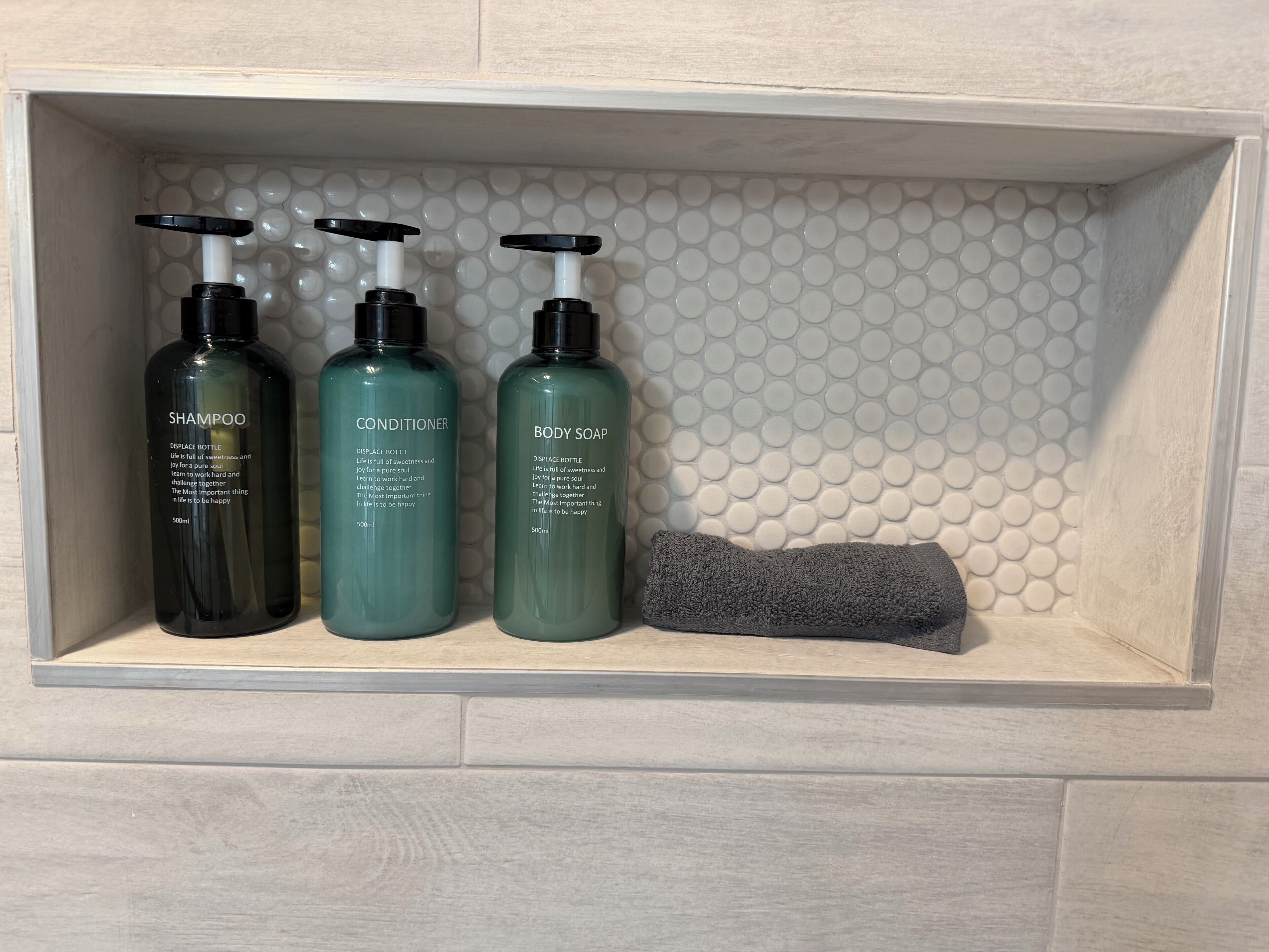En hylla i badrummet med tre flaskor schampo, balsam och duschkräm, samt en grå handduk.