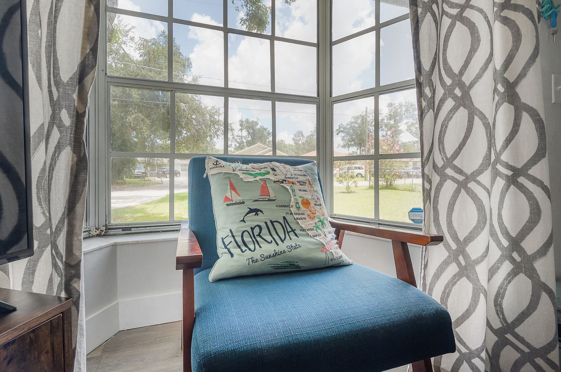 A corner room with a large window, a chair with a pillow, and a patterned curtain.