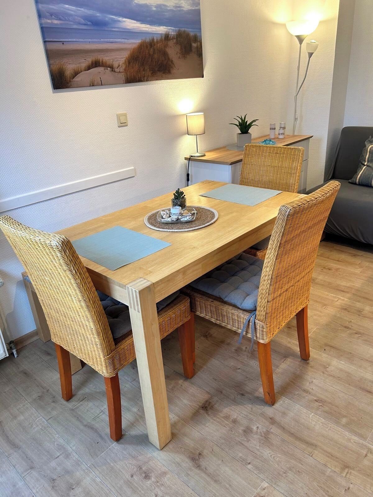 Een eethoek met een houten tafel, rieten stoelen, een lamp en een lijst met een foto van een strand.