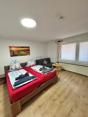 A room with two beds, a window with blinds, a radiator, a painting on the wall, and a ceiling light.