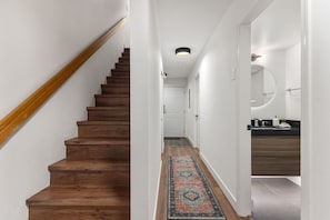 Un escalier en bois menant à une porte blanche, un couloir orné d’un tapis à motifs et une salle de bain moderne avec un plan de travail foncé.