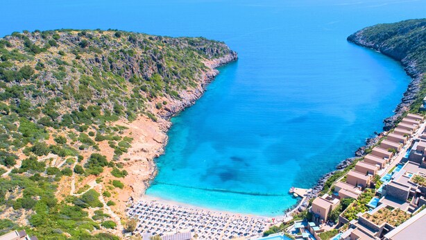 An aerial view of a coastal resort with a beach, rocky terrain, and a clear blue sea. - Cretan Lodge Heated Pool (Agios Nikolaos)