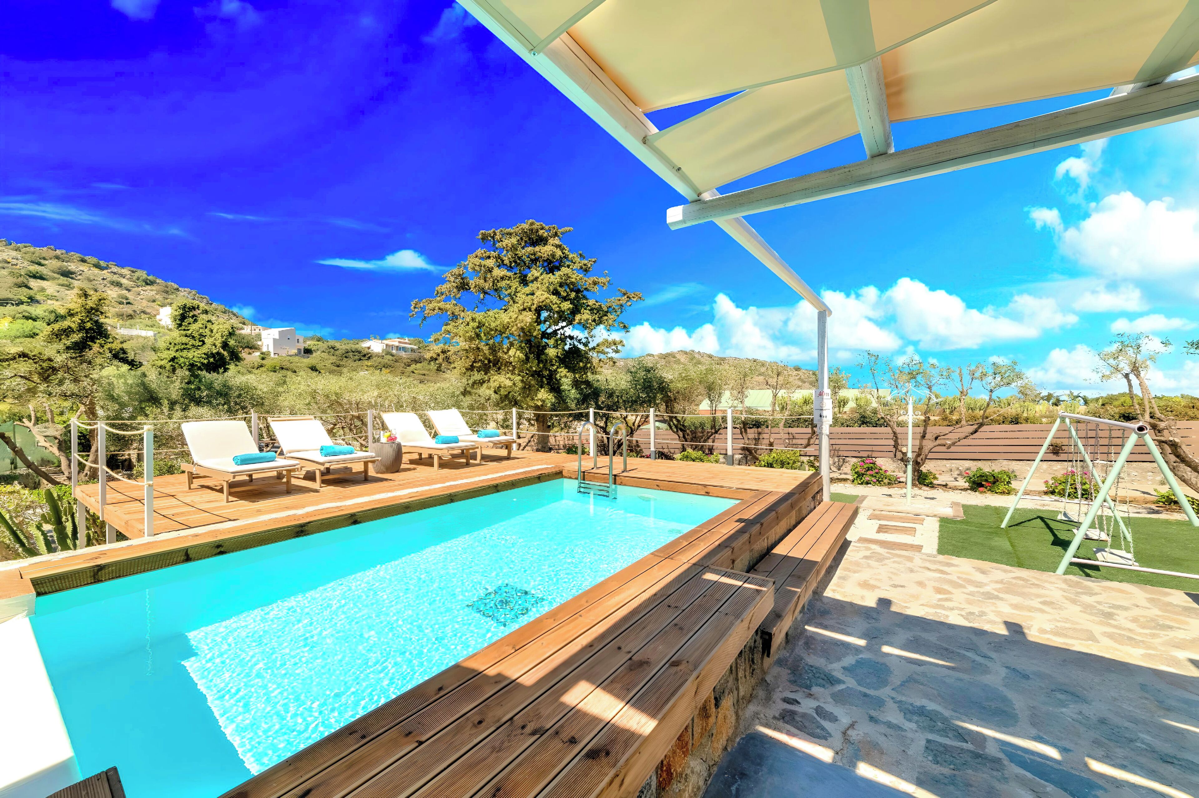 Una terraza de madera con piscina, sillas de descanso y un conjunto de columpios.