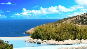 A coastal view with a clear sky, a beach, and a pool area with rocks and greenery. - Cretan Lodge Heated Pool (Agios Nikolaos)