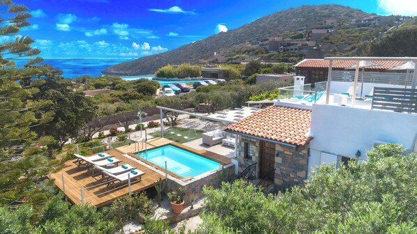 A modern villa with a swimming pool, a patio with lounge chairs, and a view of the sea and hills.