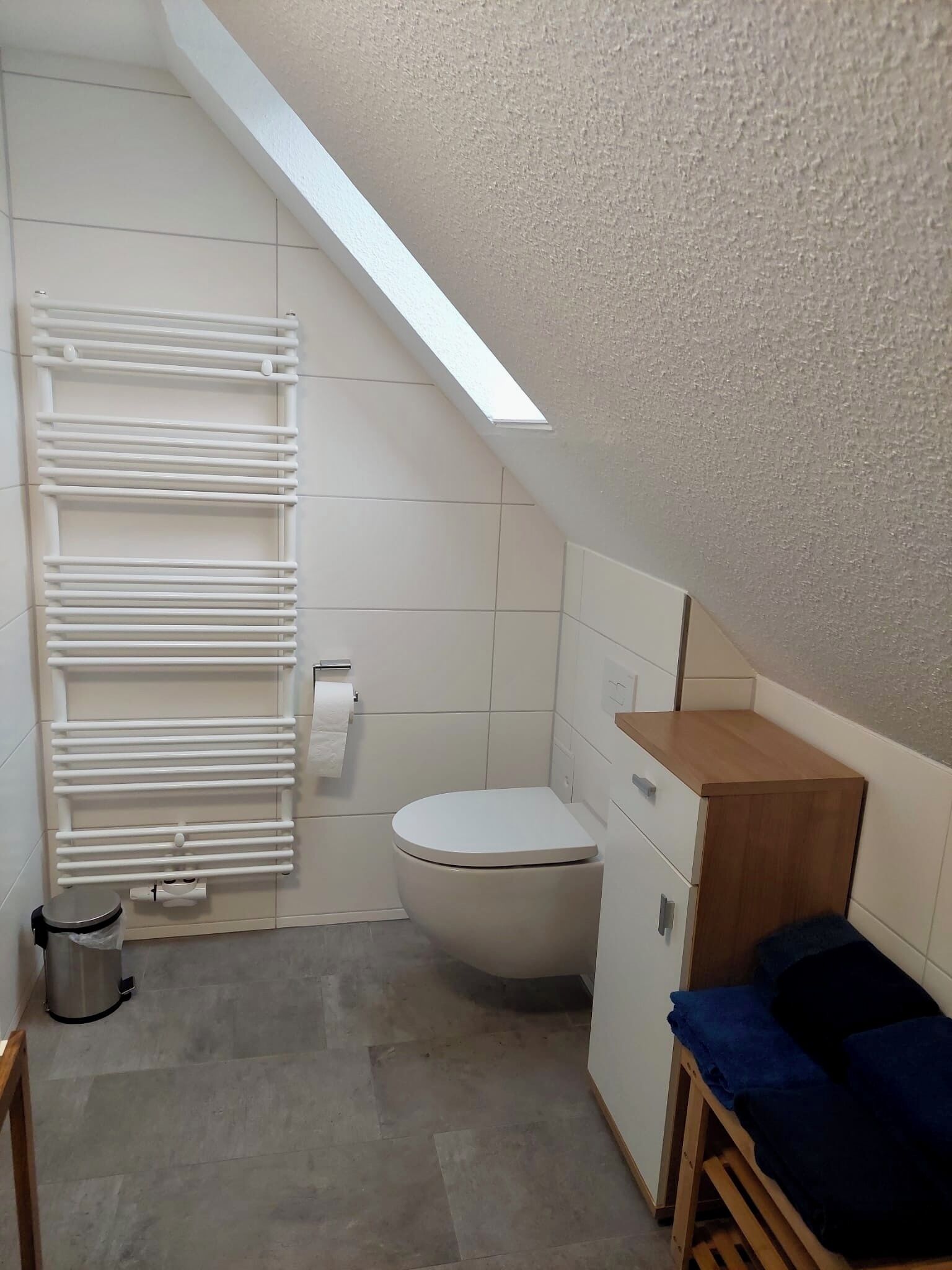 Een kleine, moderne badkamer met een wit toilet, een handdoekrek, een houten kastje en een klein houten bankje.