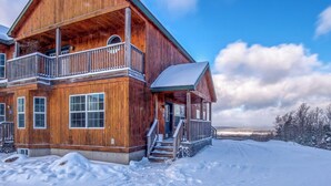A two-story wooden cabin with a balcony, a covered porch, and a snow-covered landscape.
