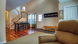 A spacious living area with a wooden staircase, a dining table with chairs, a bench, and a large window with curtains.