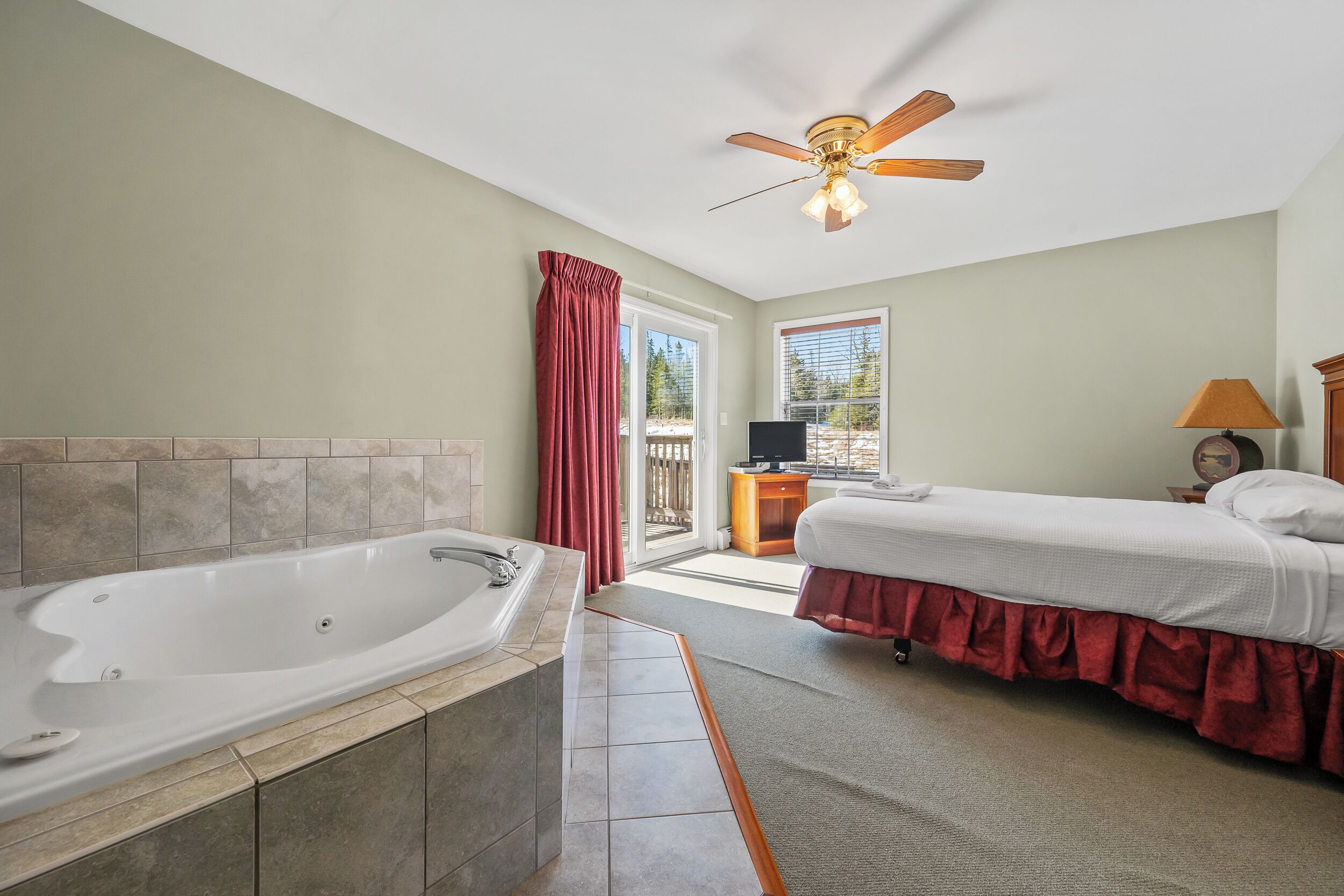 Ein Schlafzimmer mit einer großen Badewanne, einem Bett mit rotem Überwurf, einem Deckenventilator und Blick ins Freie.