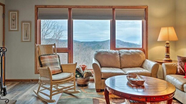 Uma sala de estar aconchegante com uma poltrona de balanço, um sofá e uma mesa de centro. Há grandes janelas com vista para o exterior.