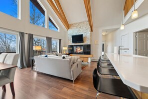 A modern living room with a fireplace, a flat-screen TV, and a dining area with bar stools. - Magalloway Loop Home 27 - Large family home at Saddleback (Rangeley)