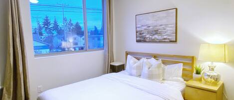 A hotel room with a large window, a bed with white linens, a bedside table with a lamp and a plant, and a painting on the wall.