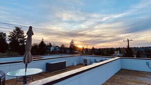 Kattoterassi, jossa on pöytä ja tuoleja, katettu aurinkovarjo ja näkymä auringonlaskuun asuinalueen yllä.