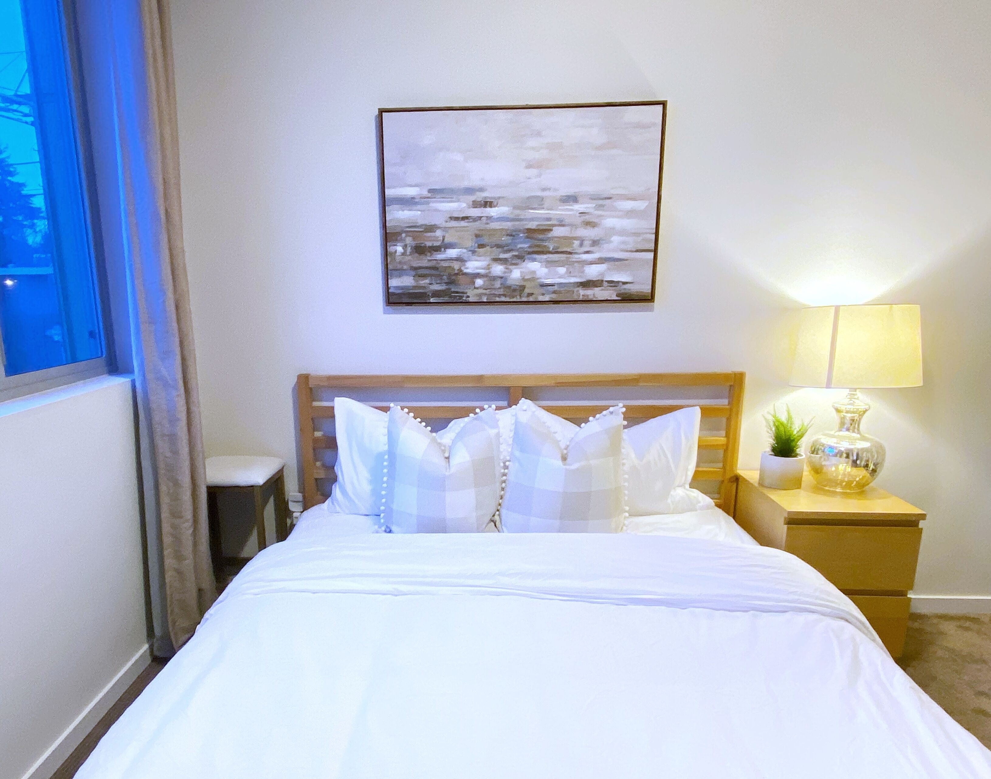 A hotel room with a bed, a nightstand with a lamp, a small plant, and a painting on the wall.