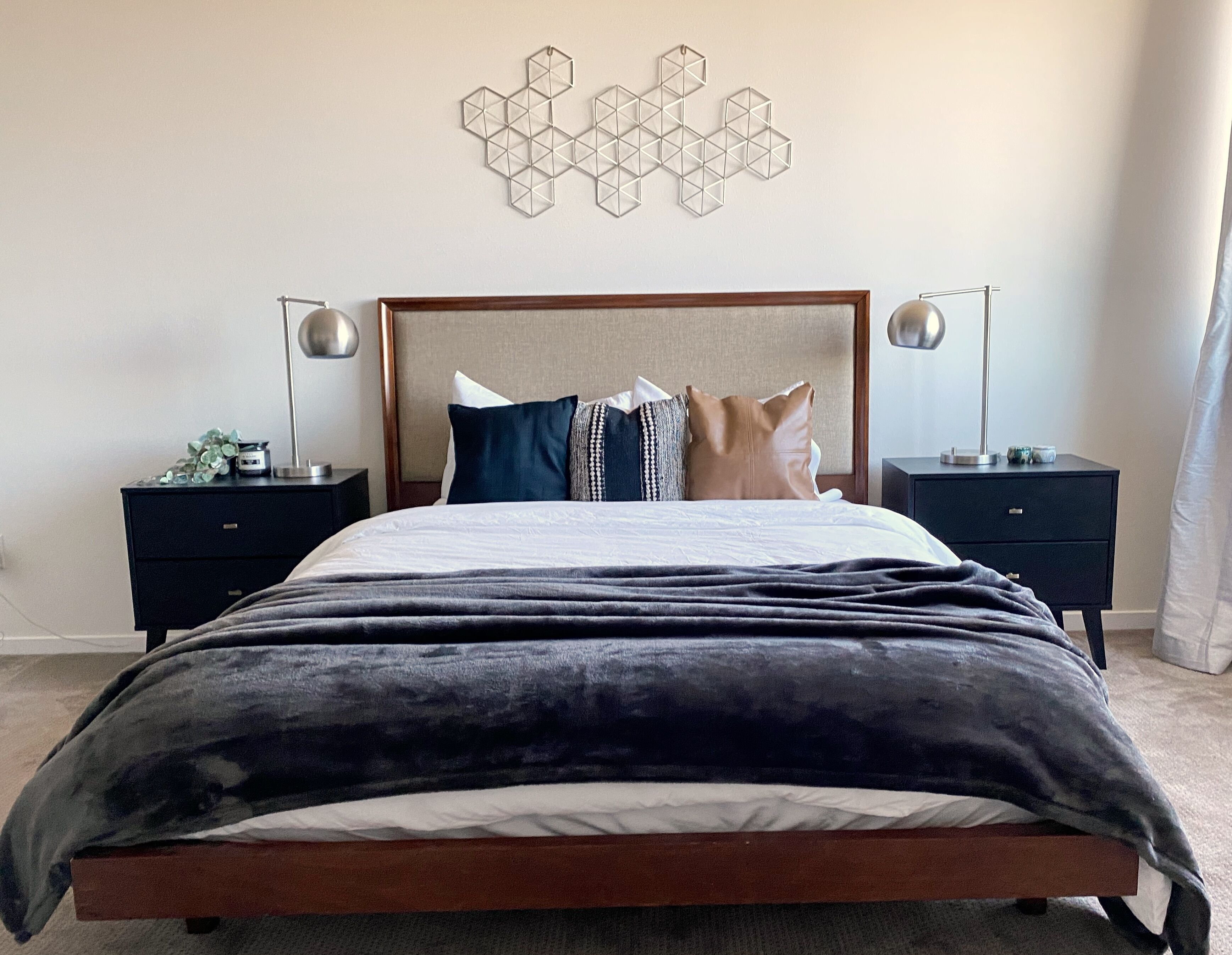 Un lit bien fait, avec une couette grise et blanche, deux tables de chevet avec des lampes et une décoration murale aux motifs géométriques.