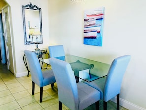 A dining area with a glass table and four blue chairs, a mirror with a decorative frame, and a painting on the wall. - Renovated Lakefront Condo · Disney 1 Mile · Fireworks Views · 2 Full Baths (Orlando)