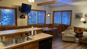A cabin-style kitchen with wooden cabinets, a dishwasher, a dining table with chairs, and large windows offering views of the forest. - Charming and Cozy, 4-Season, Fence Lake Cottage with Beautiful Sunset View (Lac du Flambeau)