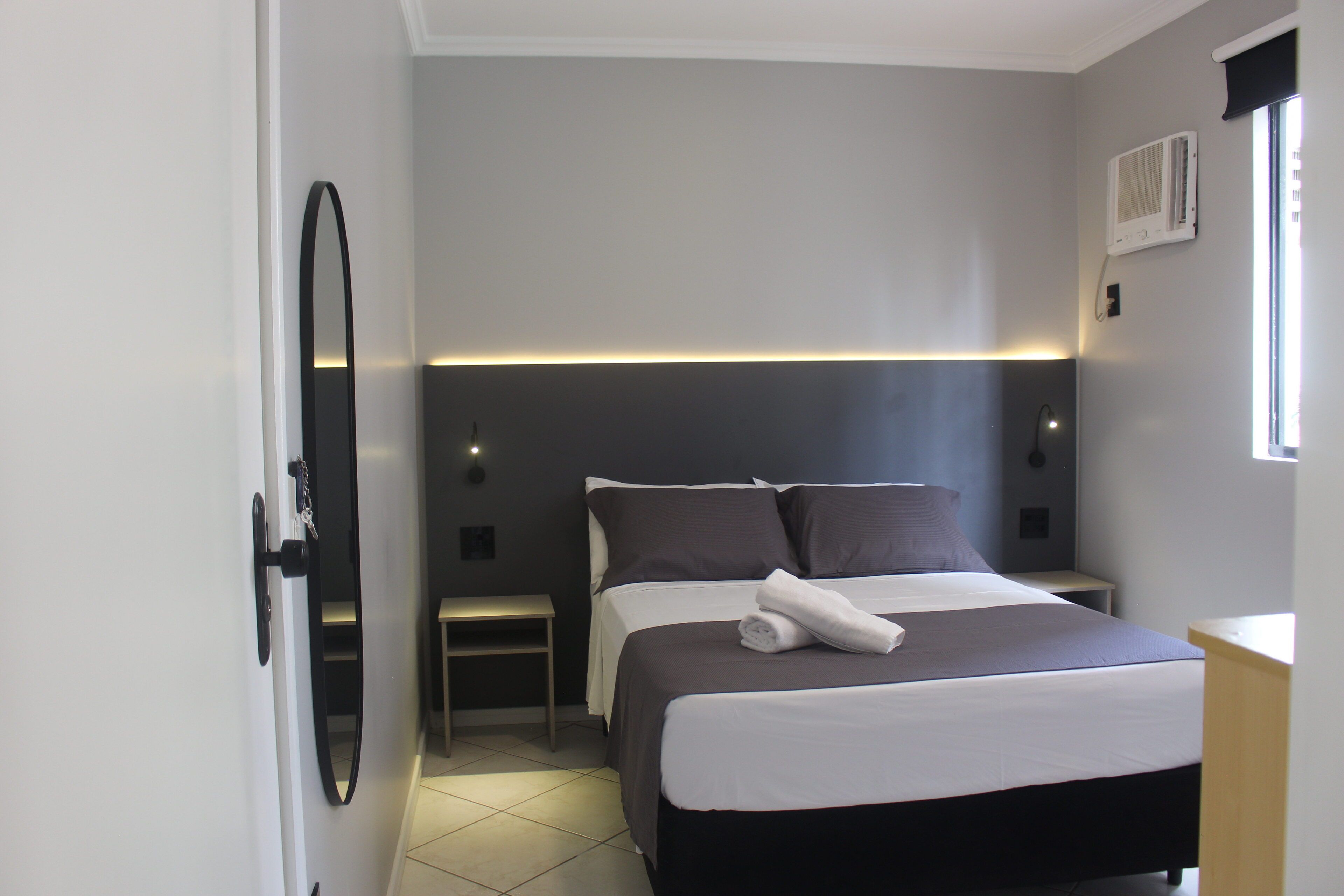 A modern hotel room with a large bed, bedside tables, a mirror, and a window with an air conditioning unit.