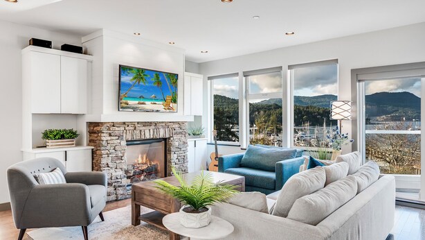 A modern living room with a stone fireplace, a sofa, a coffee table, and a view of a marina through large windows. - Bliss By the Bay (Brentwood Bay)