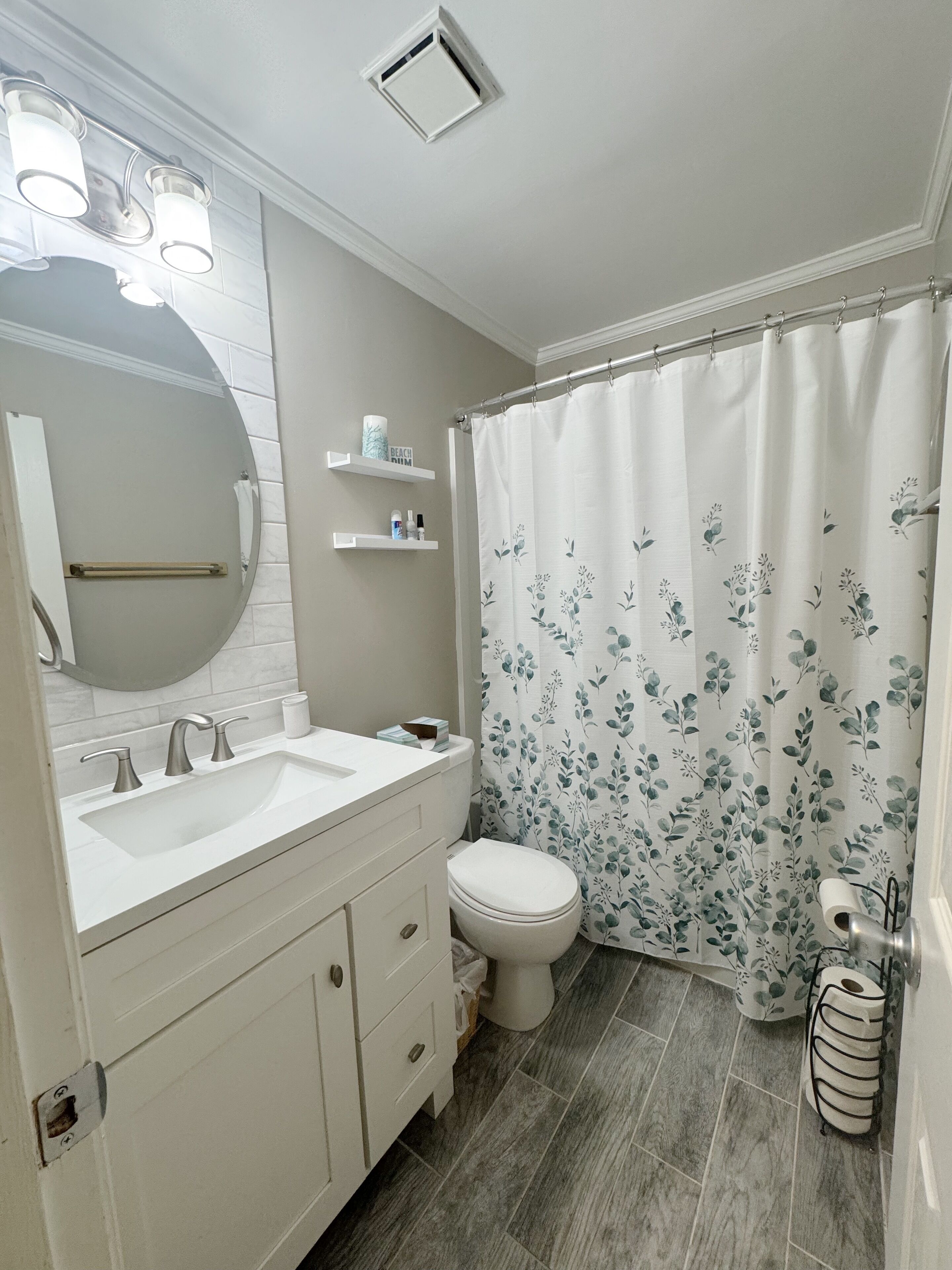 A bathroom with a white vanity, a round mirror, a toilet, and a shower curtain with a floral pattern.