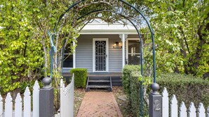 A house with a white picket fence, a brick pathway leading to the entrance, and a garden with green foliage.