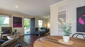 A modern living room with a wooden dining table, a sofa, and a blue couch. There is a door leading to a balcony with a view of greenery.