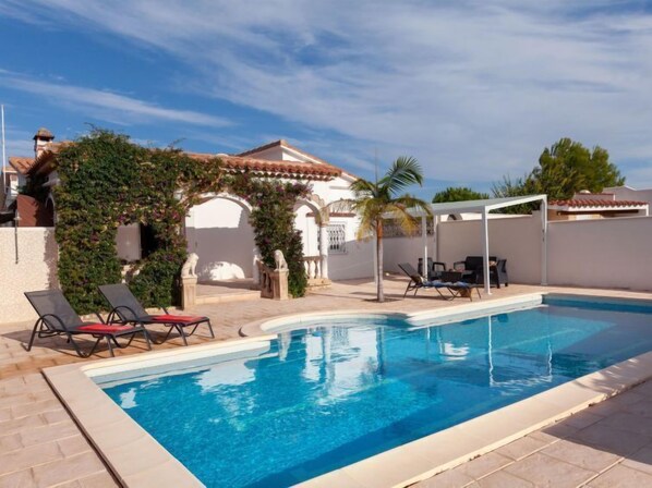 Une piscine avec des chaises longues, une pergola et un bâtiment blanc doté d’un toit en tuiles.