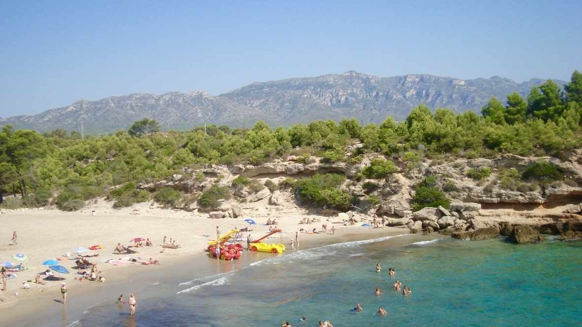 A beach with clear turquoise water, people swimming and sunbathing, and a rocky hillside with trees in the background.