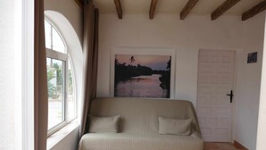 A room with a beige sofa, a large window, a door, and a painting of a sunset over water.