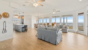 Een ruime woonkamer met een grote grijze modulaire bank, een keuken met een centraal kookeiland, en een balkon met uitzicht op de oceaan.