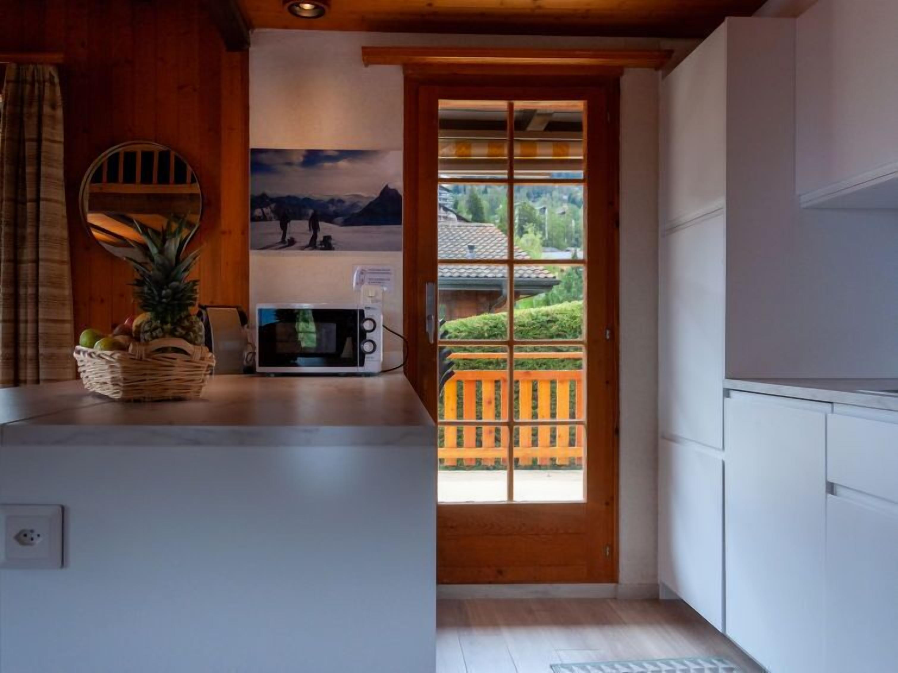 A kitchen with a microwave, a fruit basket, and a view of a wooden deck and greenery outside.