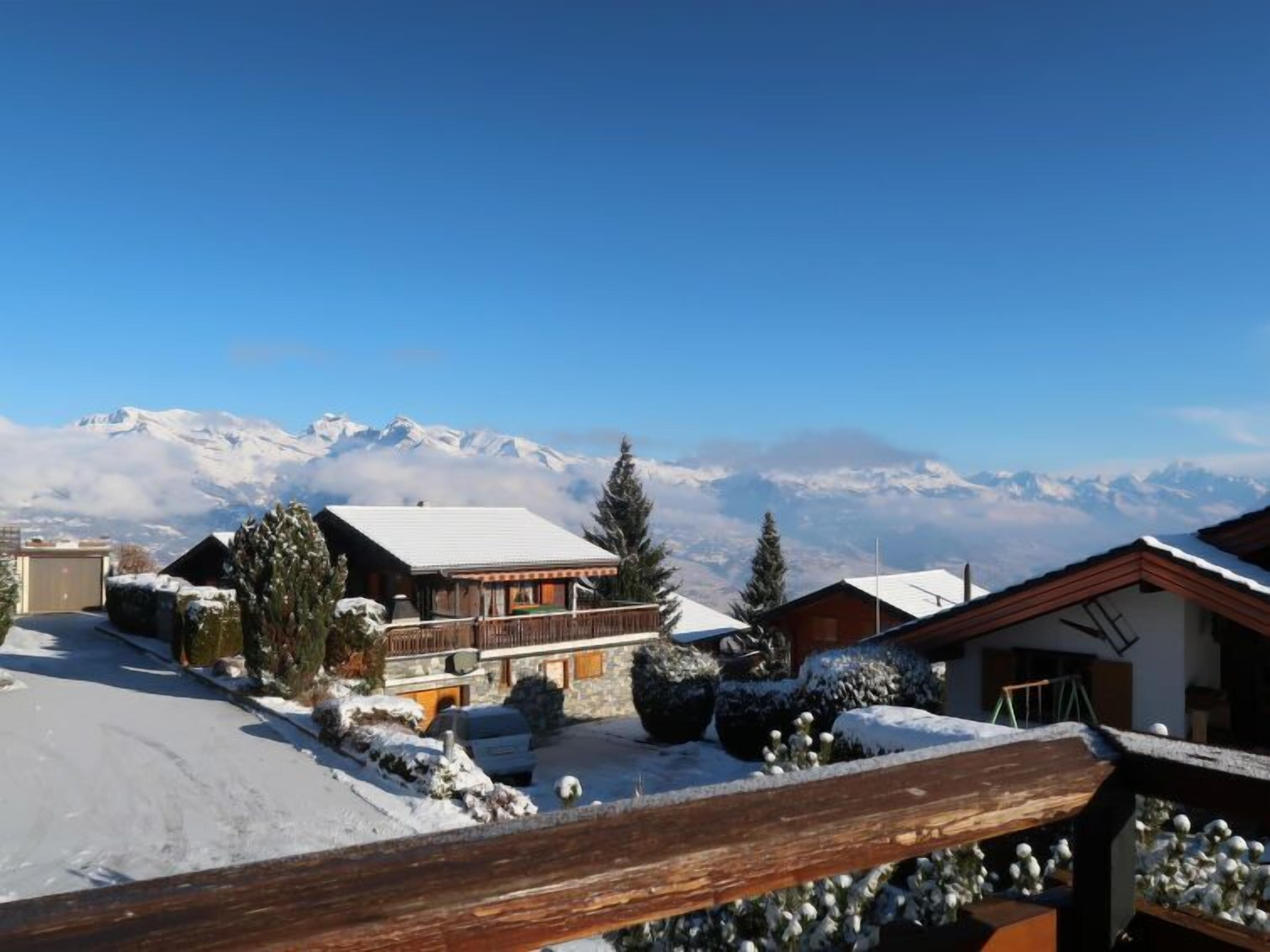 Un refugio de montaña con terraza de madera, tejados cubiertos de nieve y vistas a montañas nevadas.
